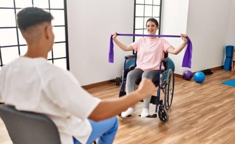 Junge Frau im Rollstuhl trainiert mit Widerstandsband in Therapie, unterstützt von einer weiteren Person, Thema Inklusion und Teilhabe
