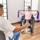 Junge Frau im Rollstuhl trainiert mit Widerstandsband in Therapie, unterstützt von einer weiteren Person, Thema Inklusion und Teilhabe