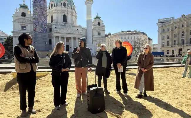 Kuratorinnen und Kuratoren sowie beteiligte Künstlerinnen und Künstler stehen im Sandbereich vor der Karlskirche bei der Klima Biennale Wien 2026