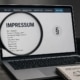 Laptop mit geöffneter Impressumsseite und Lupe als Symbol für Medienstandards, Transparenz und Aufnahmekriterien der Digitalen Allianz.
