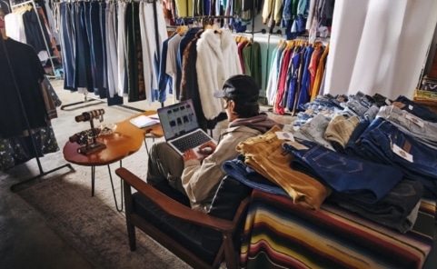Secondhand-Händler arbeitet zwischen Kleiderständern und Gebrauchtwaren an einem Laptop.