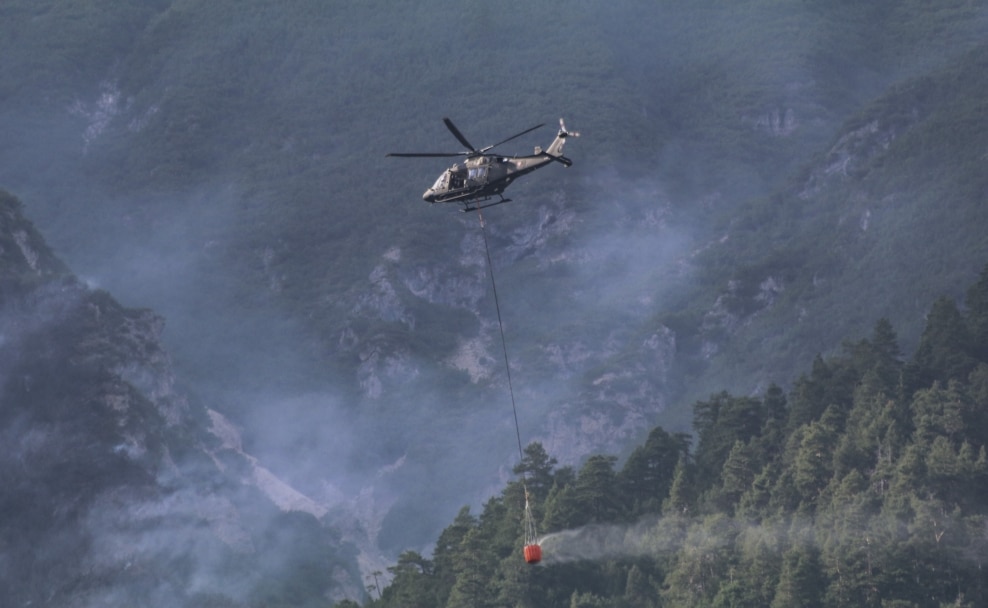 Ein AW169 Lion des Bundesheeres transportiert einen Löschwasserbehälter über bewaldetem Berggelände.