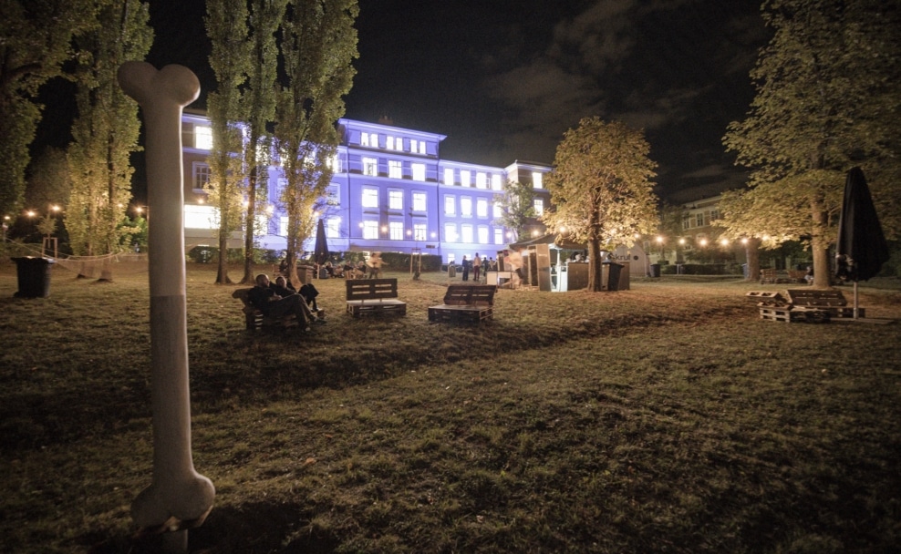 Menschen sitzen und stehen abends im Garten des beleuchteten Otto-Wagner-Areals