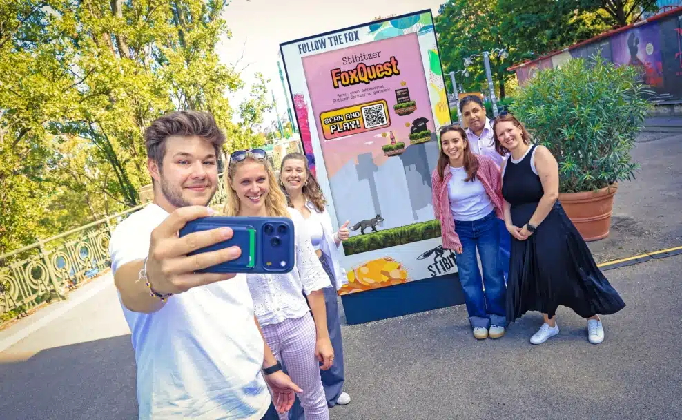 Sechs Projektbeteiligte spielen vor einem digitalen Stibitzer-FoxQuest-Screen per Smartphone.