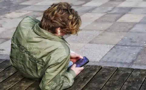 Jugendlicher sitzt mit Kopfhörern auf einer Bank und benutzt ein Smartphone.