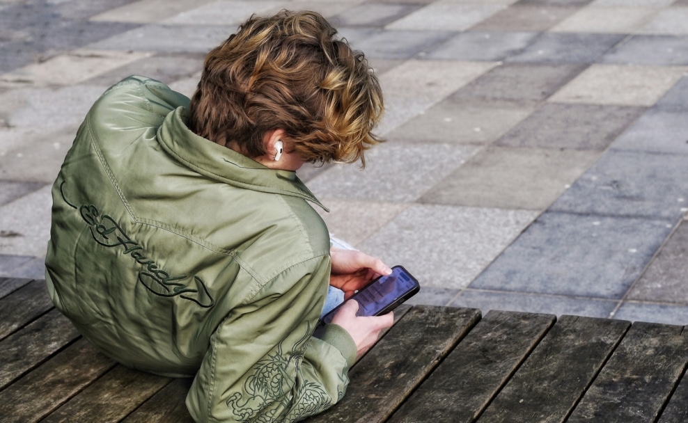 Jugendlicher sitzt mit Kopfhörern auf einer Bank und benutzt ein Smartphone.