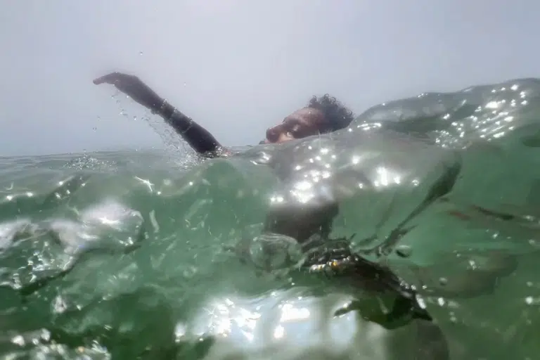 Ein Mensch schwimmt im Meer, ein Arm ragt aus dem Wasser. Eine grüne Welle verdeckt aus der tiefen Kameraperspektive einen Teil des Gesichts.