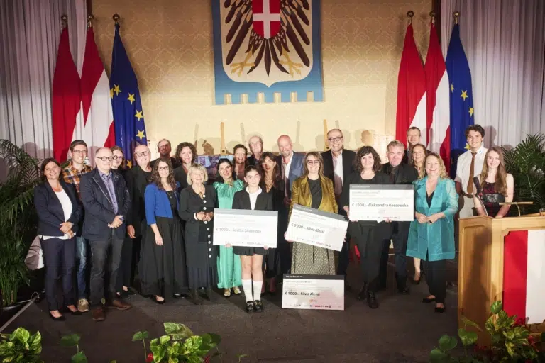 Eine große Gruppe steht im Wiener Rathaus vor österreichischen und europäischen Flaggen. Mehrere Personen halten Urkunden und große Preisschecks.