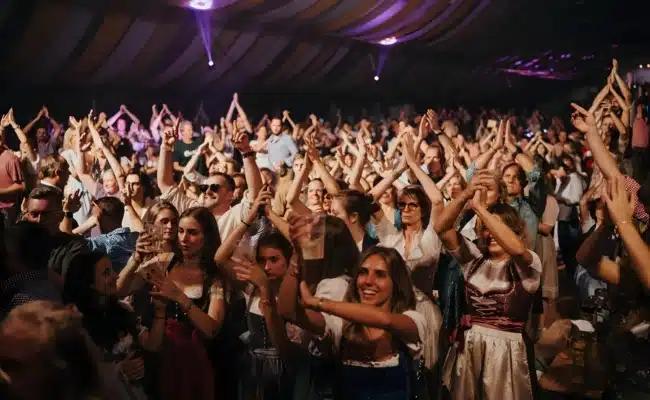 Feiernde Gäste in Dirndl und Tracht klatschen und tanzen im voll besetzten Festzelt der Brunner Wiesn.
