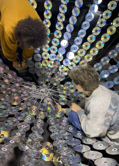 Zwei Personen arbeiten in einer raumfüllenden Installation aus Hunderten reflektierenden CDs.