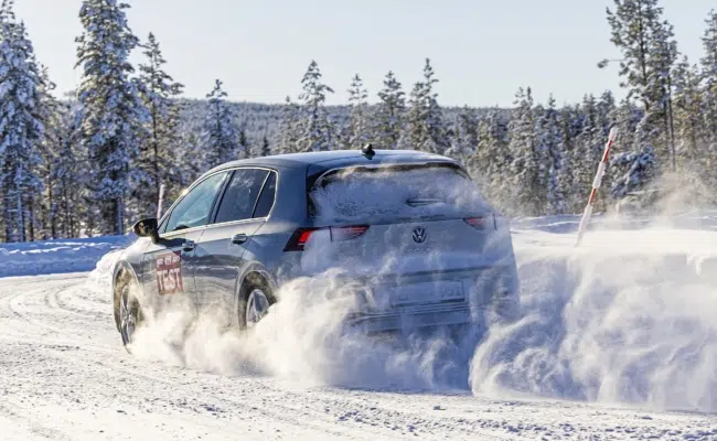 VW Golf auf einer verschneiten Teststrecke vor schneebedeckten Nadelbäumen, hinter dem Fahrzeug wirbelt Schnee auf.