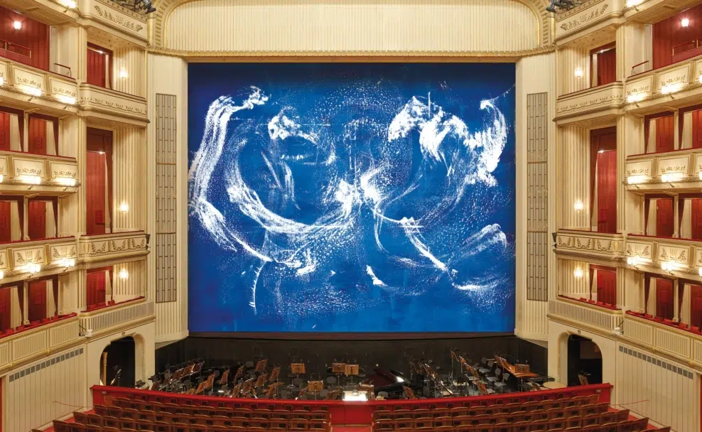 Herbert Brandls abstrakte Malerei mit weißen Spuren auf blauem Grund am Eisernen Vorhang, umrahmt vom goldenen und roten Zuschauerraum der Wiener Staatsoper.