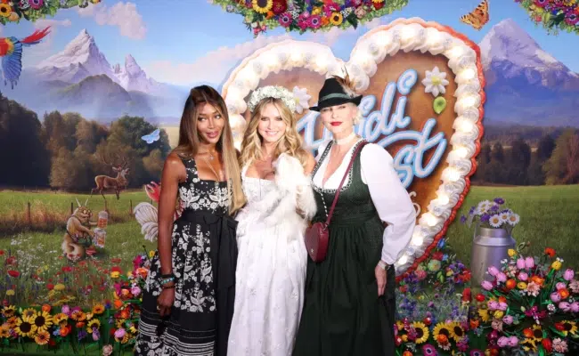 Naomi Campbell, Heidi Klum und Nadja Auermann stehen nebeneinander vor der bunten „HeidiFest“-Fotowand. Heidi hält ihren Hund im Arm.