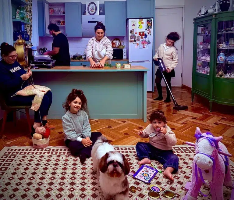 Eine Familie verteilt sich in einer bunten Wohnküche zwischen Hausarbeit und Spiel. Zwei Kinder sitzen mit Spielzeug und einem Hund auf dem Boden.