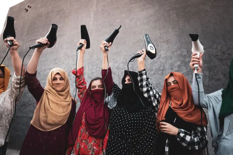 Eine Gruppe von Frauen mit Kopftüchern hält vor einer grauen Wand Haartrockner an ausgestreckten Armen in die Höhe.
