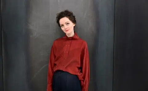 Tamara Štajner in einer rostroten Bluse und dunkler Hose vor einer grauen Wand.