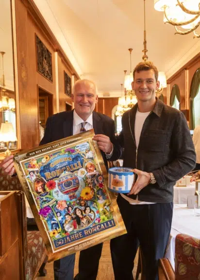 Berndt Querfeld mit gerahmtem Roncalli-Jubiläumsplakat und Ferdinand Querfeld mit der blau-weißen Gugelhupfbox im Café Landtmann.