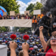 Fotorealistische KI-Collage mit Stimmabgabe, Demonstrierenden, einer Menschenmenge mit Regenschirmen sowie Polizeikräften vor Feuer und Rauch.