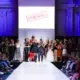 Models stehen zum Finale der Modenschau auf und neben dem Laufsteg; das Publikum applaudiert.