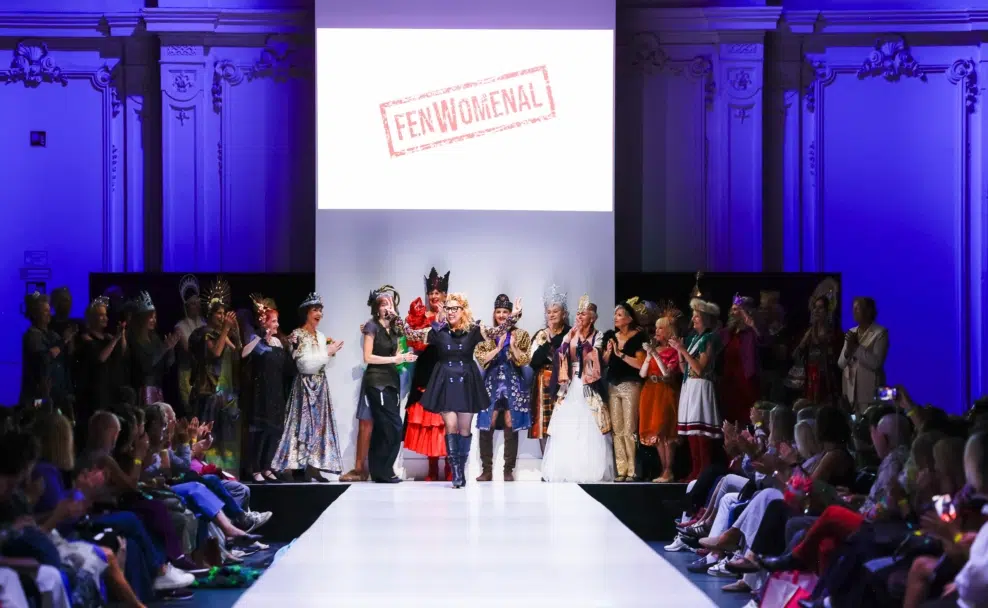 Models stehen zum Finale der Modenschau auf und neben dem Laufsteg; das Publikum applaudiert.