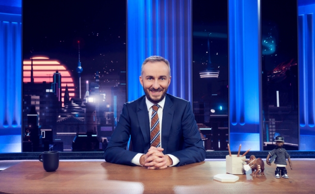 Jan Böhmermann sitzt im Studio an einem Tisch und blickt in die Kamera.