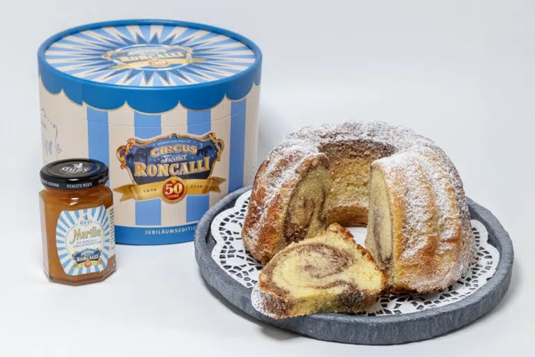 Angeschnittener Marmorgugelhupf mit Staubzucker neben der blau-weißen Roncalli-Jubiläumsbox und einem Glas STAUD’S-Marillenmarmelade.