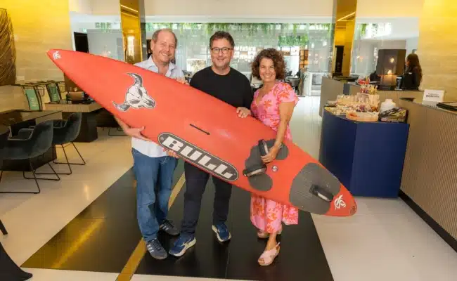 „Seesüchtig“-Team mit Surfboard Niki Klingohr, Martin Radjaby-Rasset und Sandra Klingohr halten im Le Méridien Vienna ein rotes Surfboard.