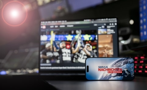 Smartphone mit „Servus Nachrichten in 90 Sekunden“ vor einem Bildschirm im ServusTV-Sendestudio