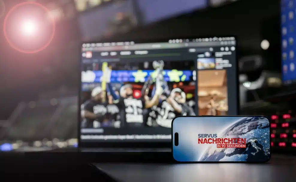 Smartphone mit „Servus Nachrichten in 90 Sekunden“ vor einem Bildschirm im ServusTV-Sendestudio