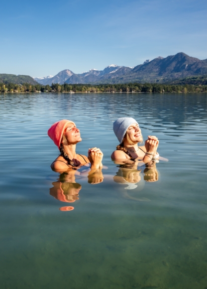 Zwei Frauen mit Mützen im Faaker See, dahinter das Ufer und eine Bergkette.