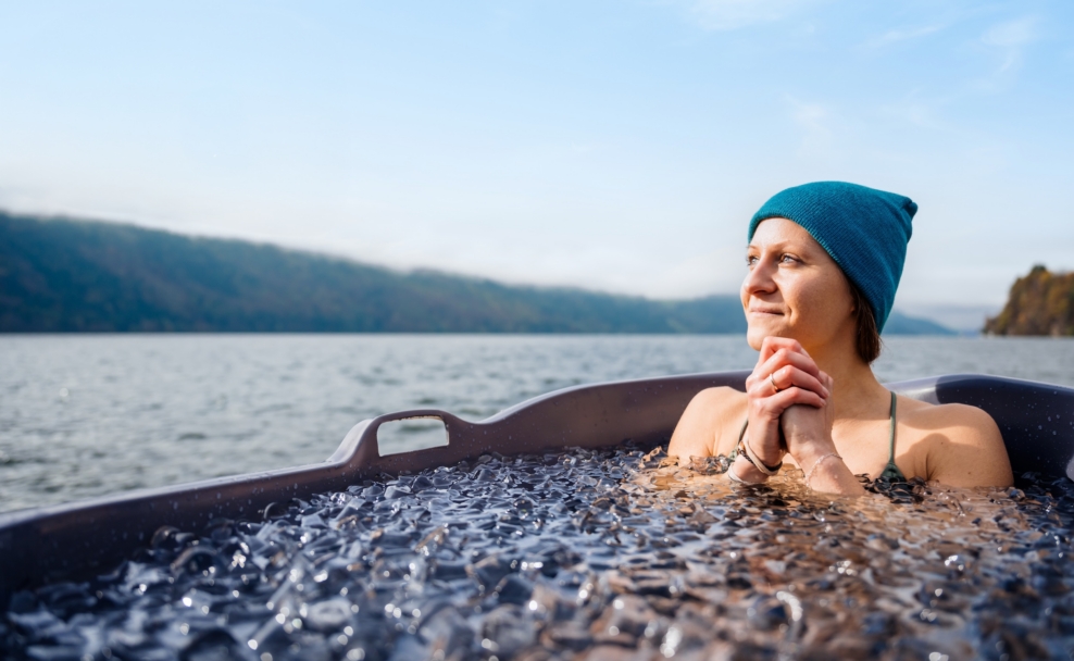 Eine Frau mit blauer Mütze sitzt in einer Wanne mit Eisstücken vor dem Millstätter See.