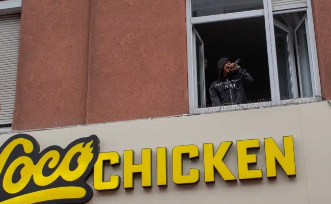 Luciano spricht mit einem Mikrofon aus dem offenen Fenster direkt über dem gelben Loco-Chicken-Schriftzug zu den Fans.