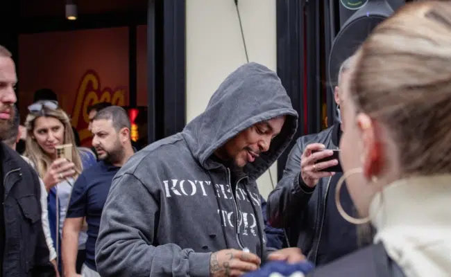 Luciano mit hochgezogener Kapuze gibt vor Loco Chicken ein Autogramm, umringt von Fans.
