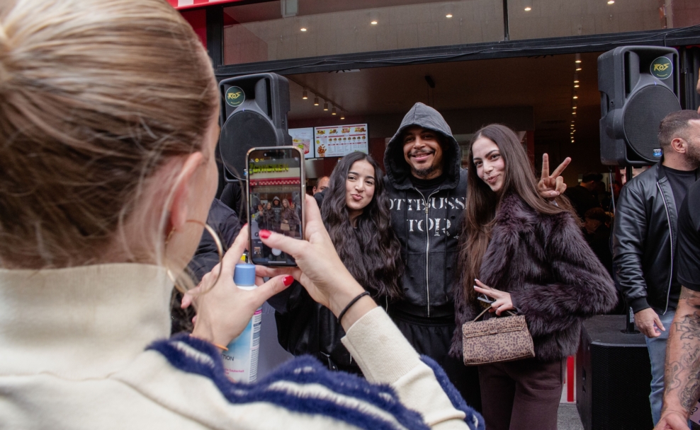 Luciano posiert mit zwei Fans am Eingang von Loco Chicken, eine Person im Vordergrund fotografiert mit dem Smartphone.