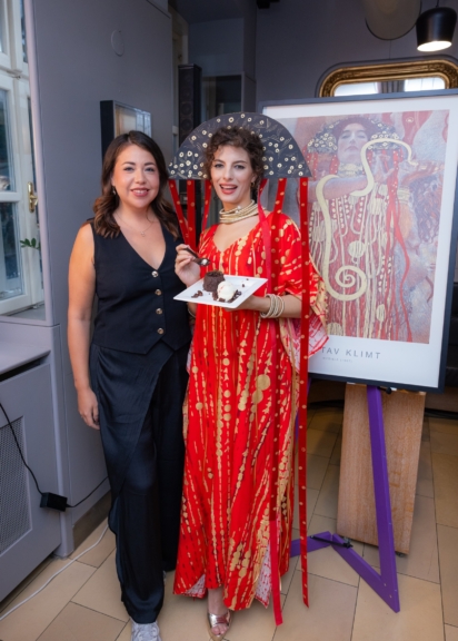 Mariella Alexandra Chaparro steht links neben Enya Rock im rot-goldenen Hygieia-Kostüm vor einer Klimt-Reproduktion.