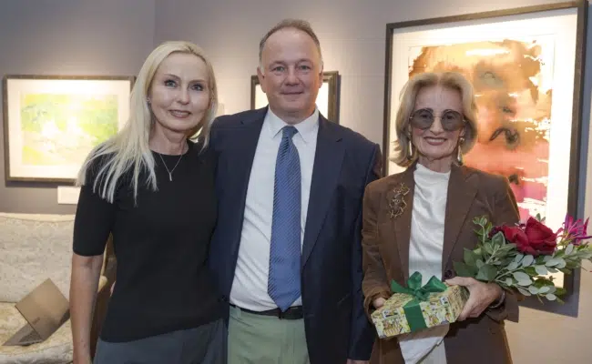 Monika Pelz, Wolfgang Pelz und Elisabeth Gürtler stehen vor ausgestellten Gemälden. Gürtler hält Blumen und ein Geschenk.