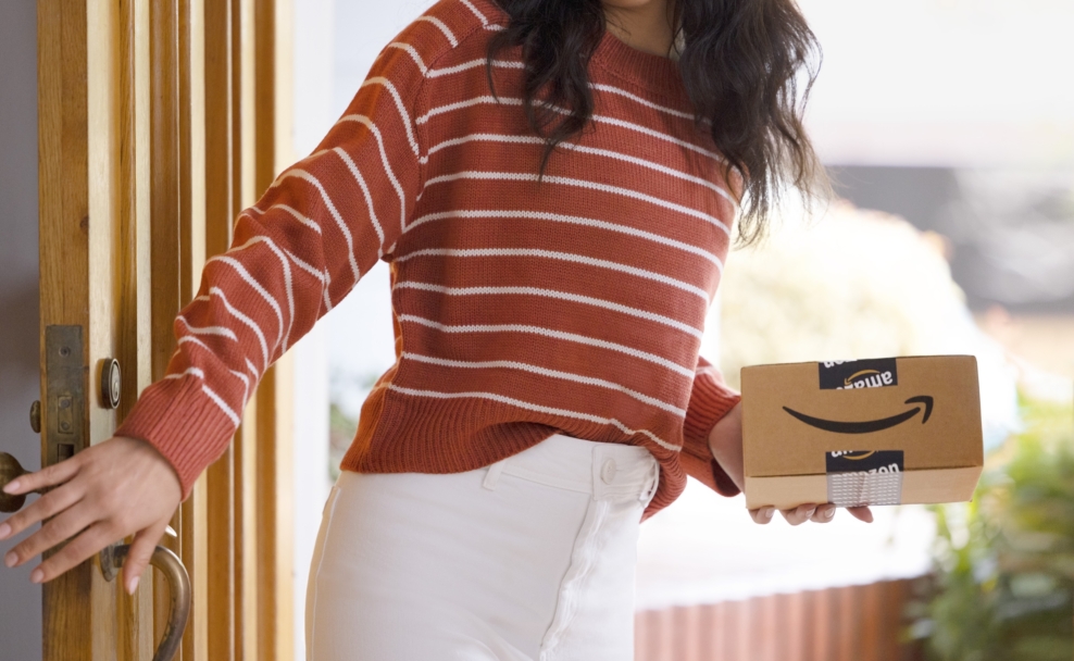 Eine Person im gestreiften Pullover steht mit einem Amazon-Paket an einer geöffneten Haustür.