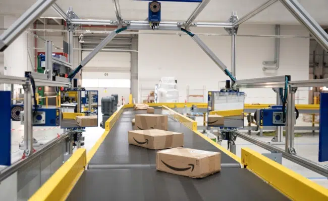 Mehrere Amazon-Pakete liegen auf einem Förderband zwischen gelben Begrenzungen in einer Logistikanlage.
