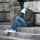 Person in grüner Winterjacke und Jeans sitzt mit gesenktem Kopf auf einer Steintreppe vor einer Steinwand.