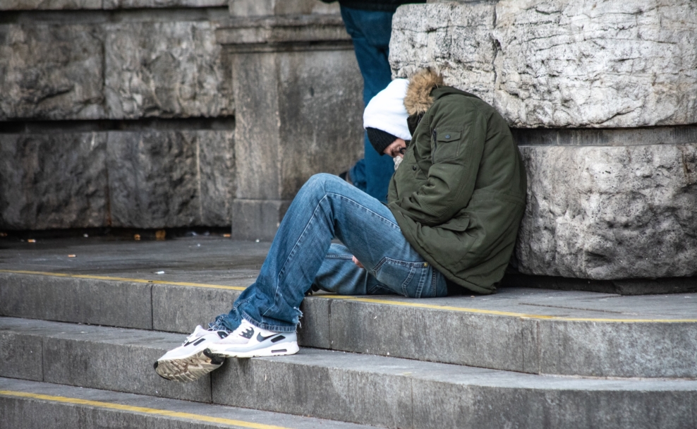 Person in grüner Winterjacke und Jeans sitzt mit gesenktem Kopf auf einer Steintreppe vor einer Steinwand.
