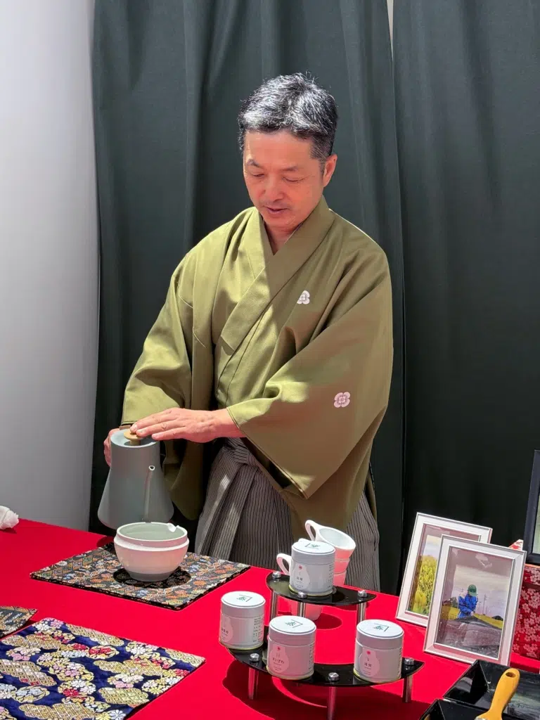 Koshi Hagimura im grünen Kimono gießt Wasser aus einer grauen Kanne in eine Teeschale auf einem roten Tisch.